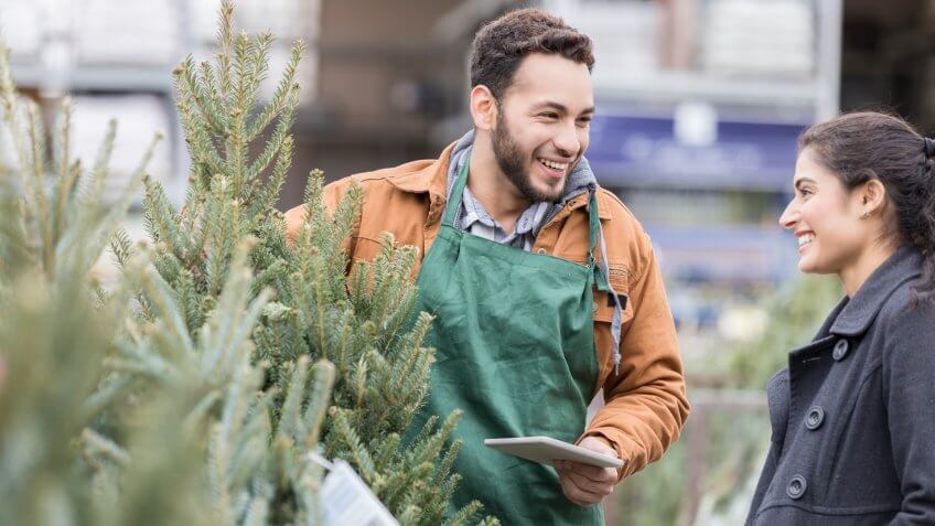 Confident male Christmas tree lot owner helps a female customer pick out a Christmas tree for her home.