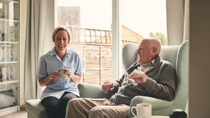 Indoor shot of smiling senior man and female carer enjoying coffee in living room.