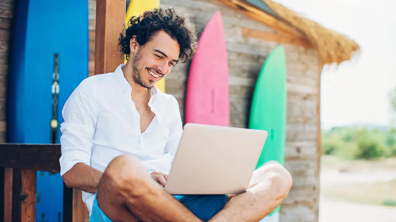 Cheerful young man using laptop with surfing boards at the background.