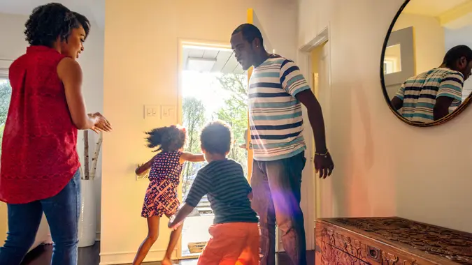 Parents are standing in the hall as their children are running towards an open front door in excitement.