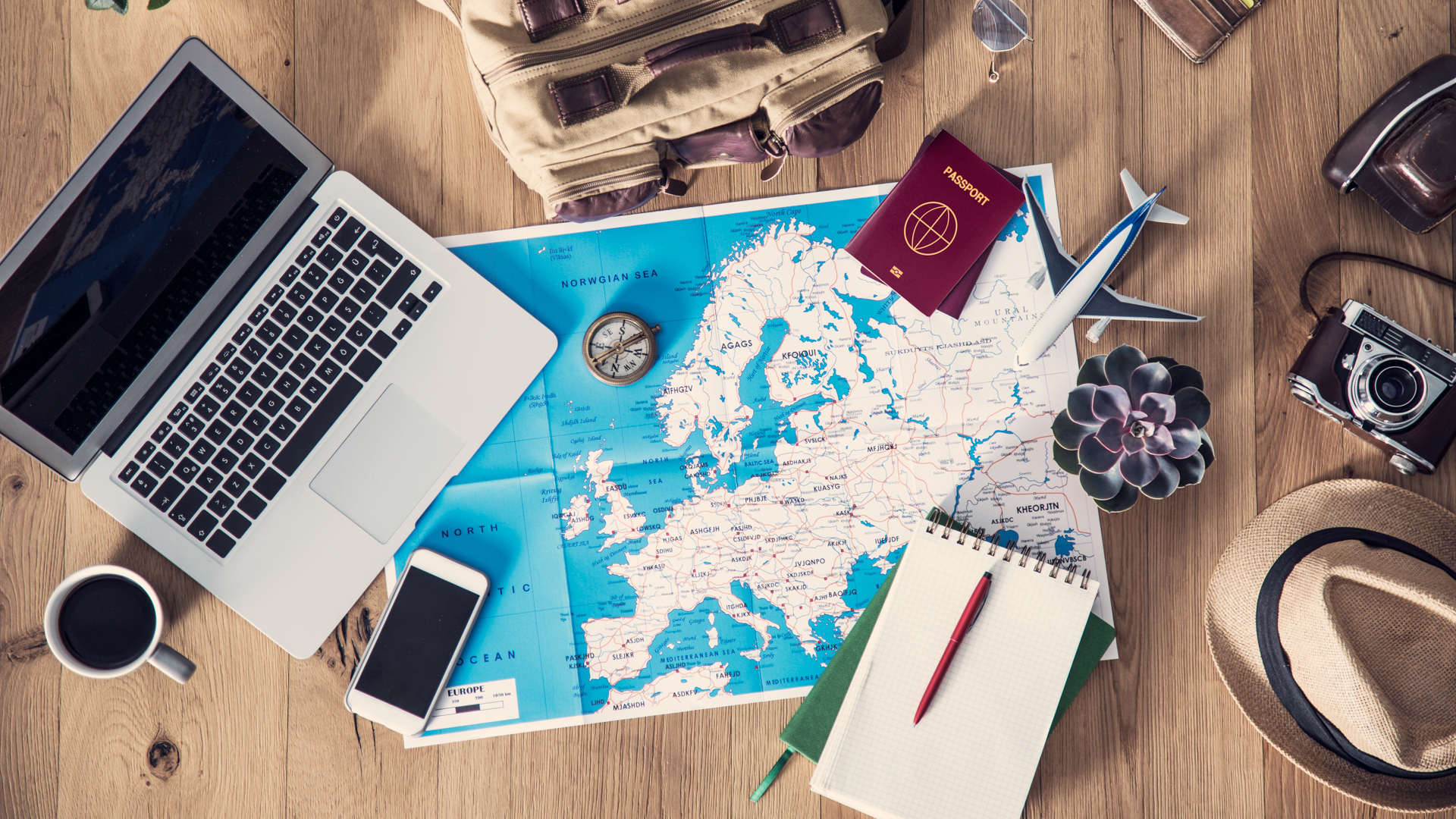  A wooden table with a map, a laptop, a camera, a passport, a compass, a notebook, a pen, a coffee cup, a backpack, a hat, a succulent, and a toy airplane.