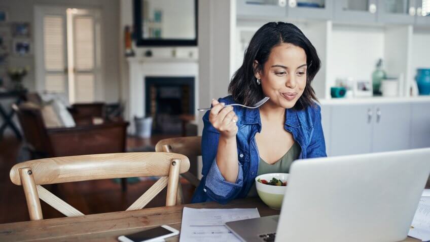 Shot of a young woman using a laptop and having a salad while working from home.