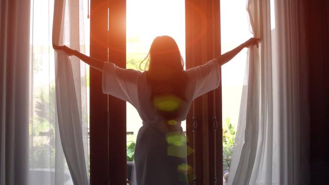 Young woman in bathrobe open curtains and stretch standing near the window at home.