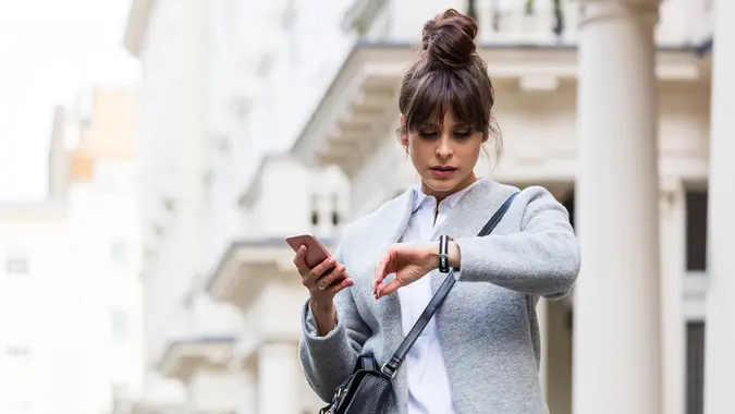 Elegant worried woman standing in front of city house in London, holding smart phone in hands and checking time.
