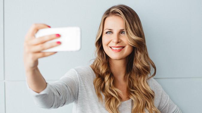 Beautiful mid-age woman standing by blue wall in office or home, using smart phone to take selfies.