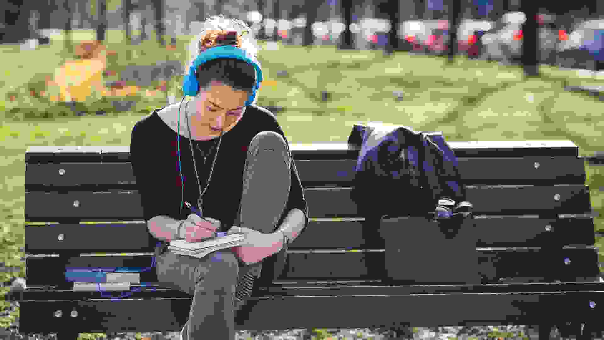 Student girl reading on a bench in the park and listening to music.