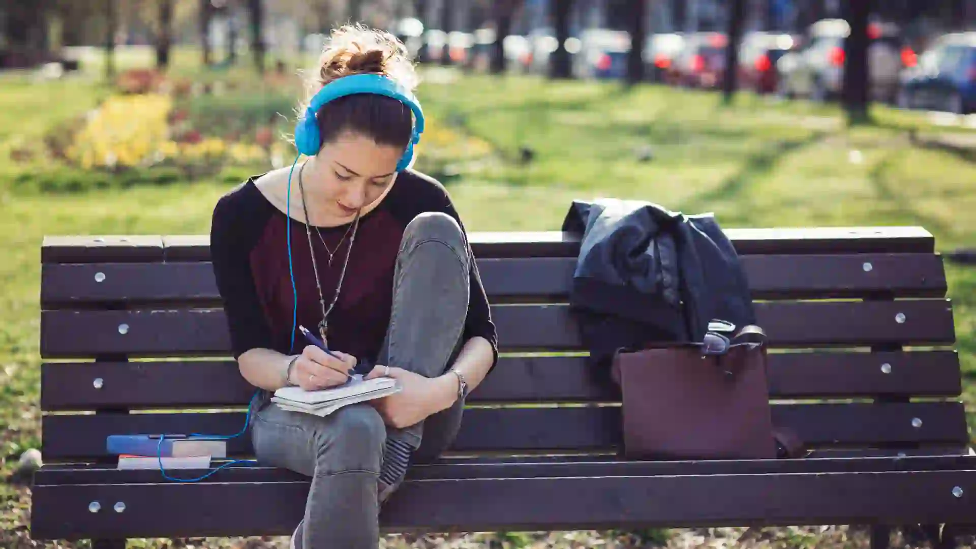 Student girl reading on a bench in the park and listening to music.
