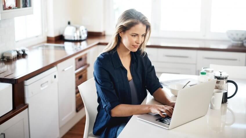 Shot of a young woman using a laptop while having breakfast in the kitchen at home.
