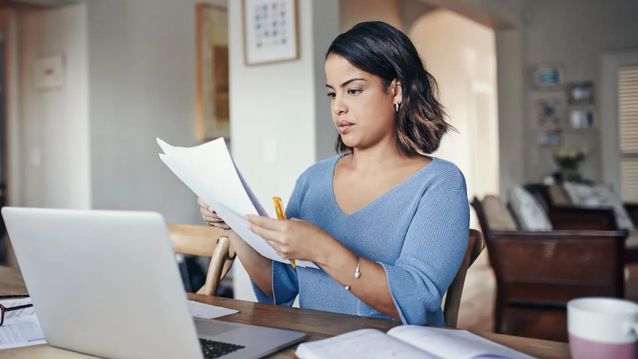 Shot of a young woman using a laptop and  going through paperwork while working from home.