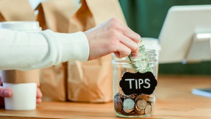 Unrecognizable coffee shop customer inserts cash into a tip jar at the checkout counter.