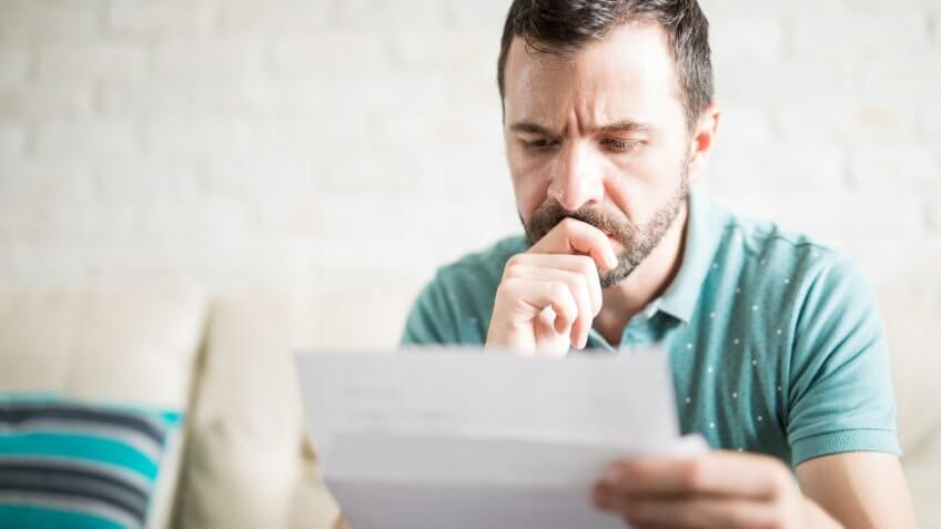 Close up shot of a young adult man reading the mail in the living room and worried about his credit card payments.