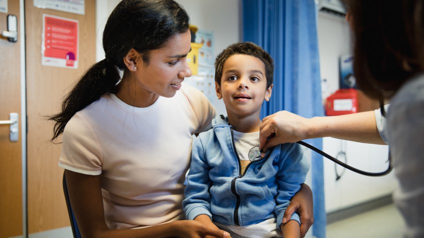 Doctor uses a stethoscope on a young boy who is sitting on his mother's knee.