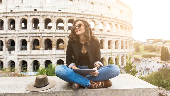 Woman sitting legs crossed in front of Coliseum and resting, using digital tablet.