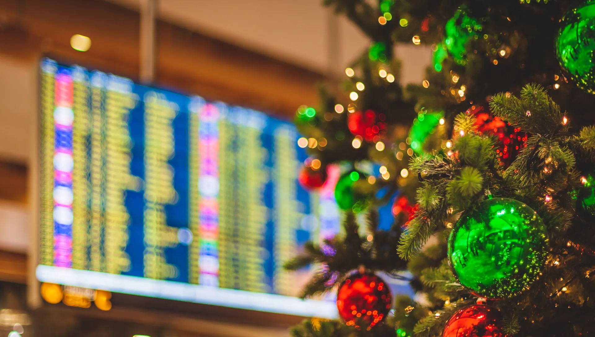 An unrecognizable airport location with a decorated Christmas Tree in the foreground an a flight departures display visible in the background.