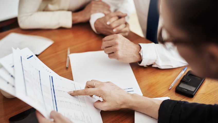 Over the shoulder shot of an unrecognizable couple speaking with a tax preparer around a table inside of a building during the day.