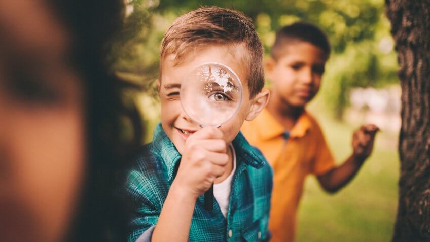 Portrait of a little boy holding up a large round magnifying glass to his face, making his eye look humourously large, while playing with friends in a summer park.