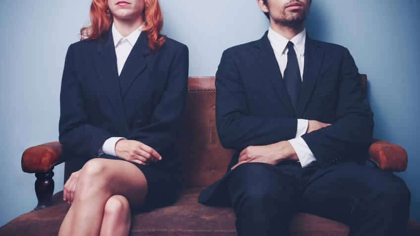 Two sharply dressed business people are waiting on a sofa with a serious expression on their faces.