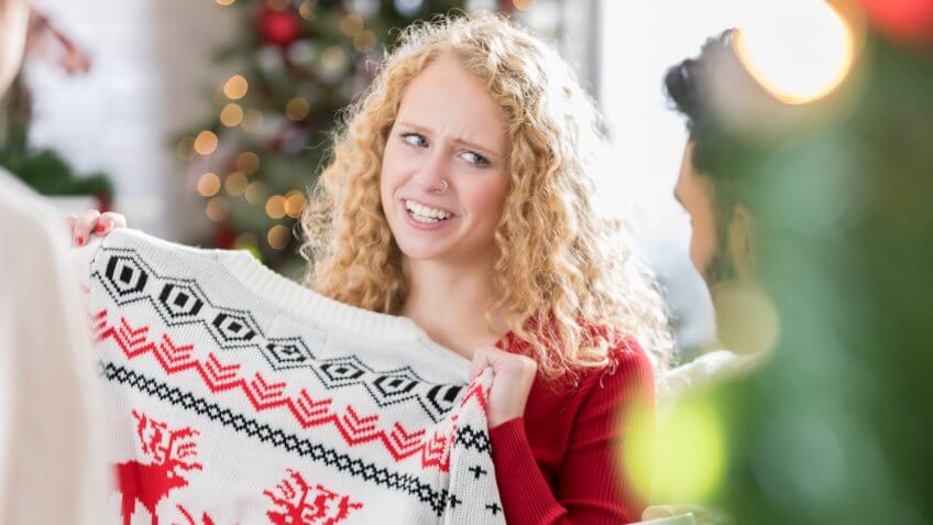 Young Caucasian woman makes a face while holding an ugly Christmas sweater.