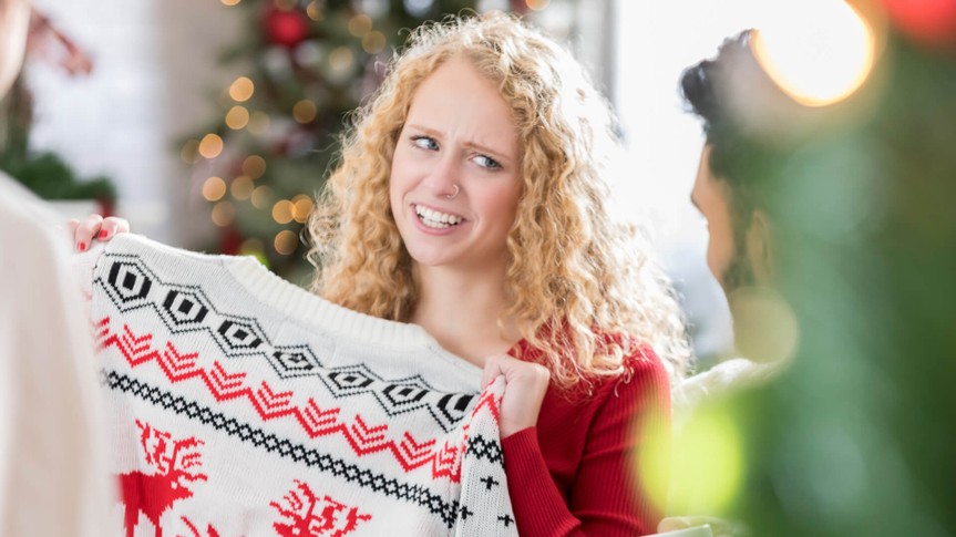 Young Caucasian woman makes a face while holding an ugly Christmas sweater.