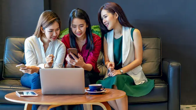 Group of charming beautiful Asian women using smartphone and laptop, chatting on sofa at cafe, modern lifestyle with gadget technology or working woman on casual business concept.