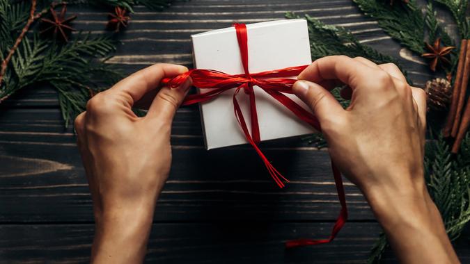 hands wrapping christmas present with red ribbon on stylish wooden background flat lay with green branches.