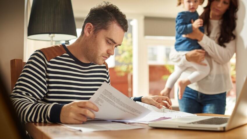 A young husband and father sits at a dining table looking through the household bills and looks worried .