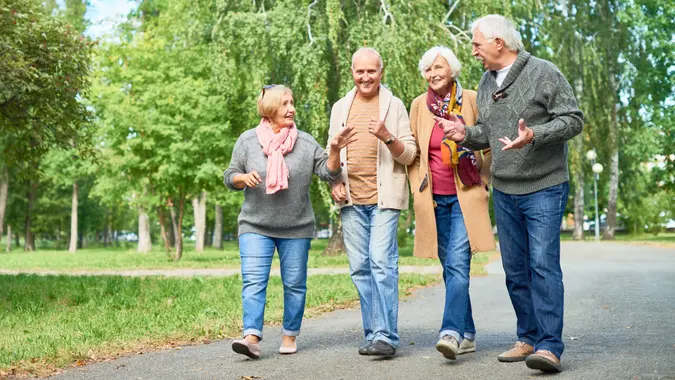 Joyful group of senior friends wearing warm clothes walking along park alley and chatting animatedly with each other, picturesque view on background.