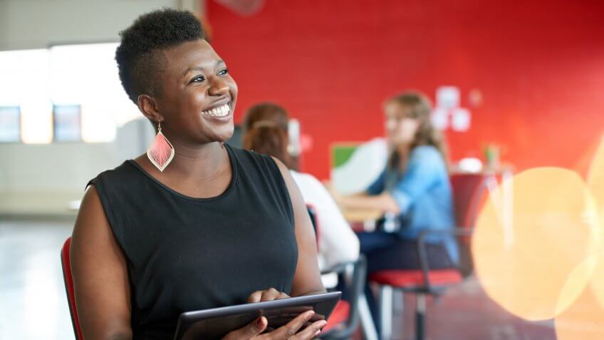 Casual portrait of an african american business woman using technology in a bright and sunny startup with the team in the background.