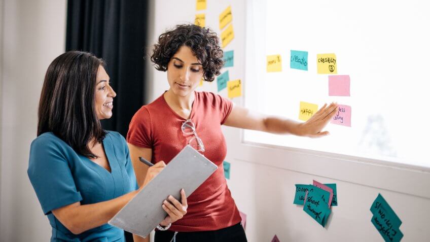 Two businesswomen are preparing presentation of project results and putting sticky notes on the wall in the seminar room.
