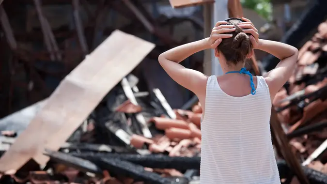 woman standing in front of burned out house and holding her head with both hands.