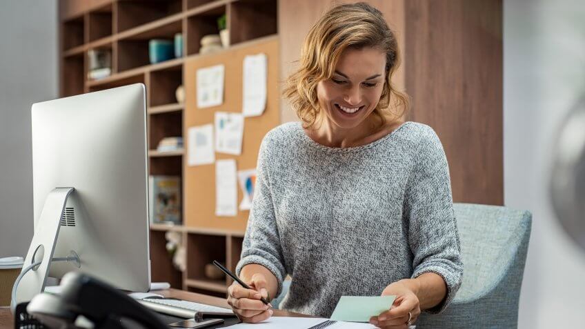 Smiling busy employee in casual reading notes and writing on notebook.