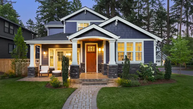 facade of home with manicured lawn, landscaping, and backdrop of trees and dark blue sky.