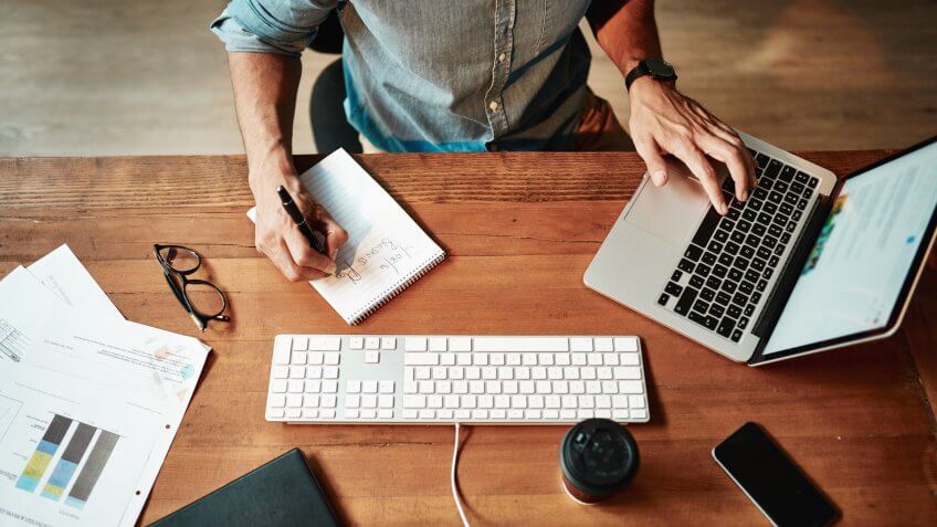 Cropped high angle shot of a businessman using a laptop and writing notes at his desk in a modern office.