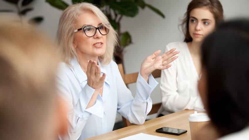 Attractive aged businesswoman, teacher or mentor coach speaking to young people, senior woman in glasses teaching audience at training seminar, female business leader speaker talking at meeting.
