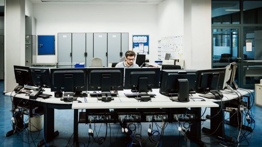 Engineer is working behind several work stations in a big control room.