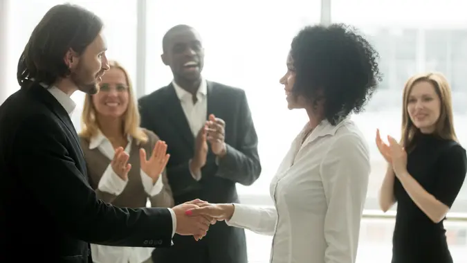 Grateful boss handshaking promoting african businesswoman congratulating with career achievement while colleagues applauding cheering successful worker, appreciation handshake, employee recognition.