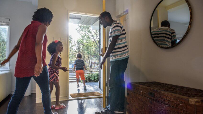 The boy is heading out through the door and the mother and father are encouraging the daughter to hurry up so they can leave the house on time.
