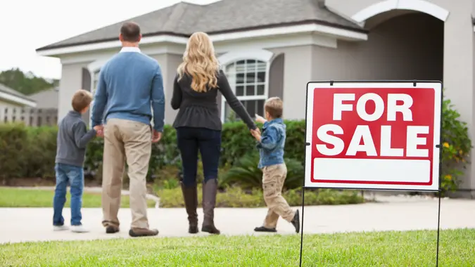 Family with two boys (4 and 6 years) standing in front of house with FOR SALE sign in front yard.