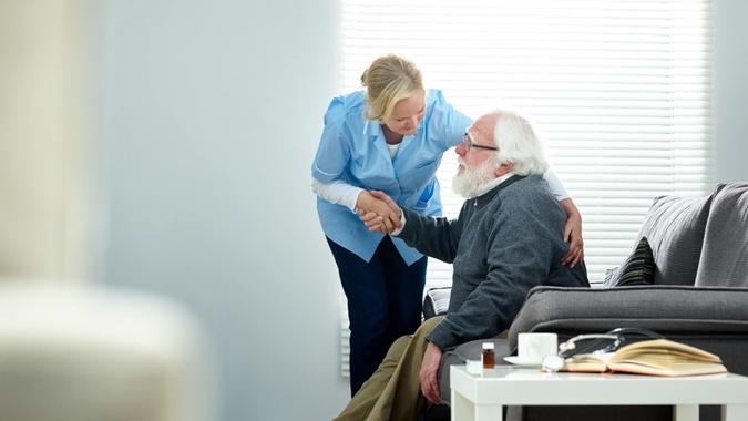 Female caregiver helping senior man get up from couch in living room - Indoors.