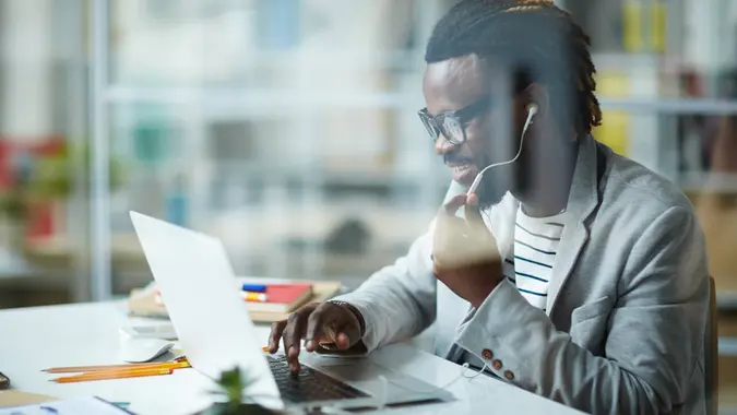 Modern young African-American entrepreneur working at successful start-up business in small office, talking with partners using phone headset and laptop, behind glass wall shot.