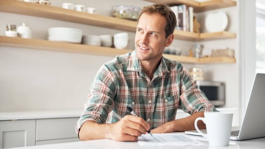 Shot of a mature man writing notes while using a laptop at home.