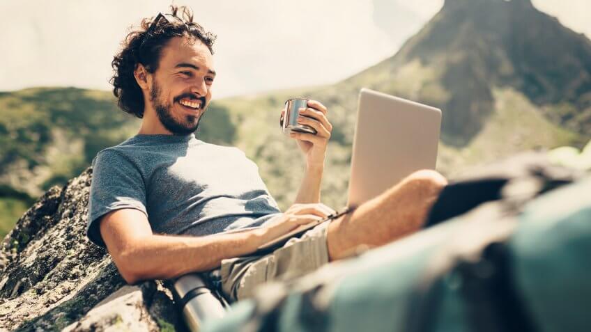 Male hiker having a drink while he's using a laptop on a rock near a lake in the mountain.