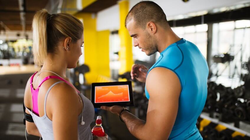 Young sporty woman is discussing workout plan, progress and statistics with her fitness instructor using digital tablet.