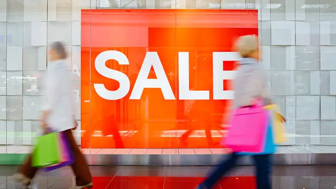 Large orange "Sale" sign with shoppers holding shopping bags walking past it.