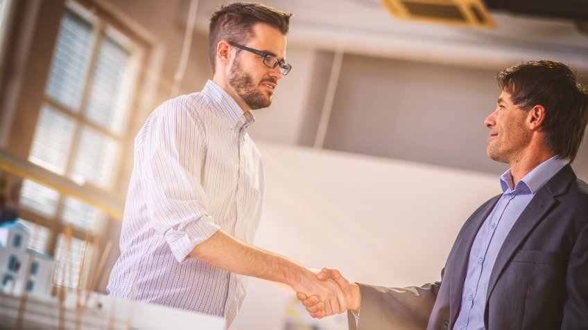 Architect shaking hands with his client while standing beside a desk with an architectural model.