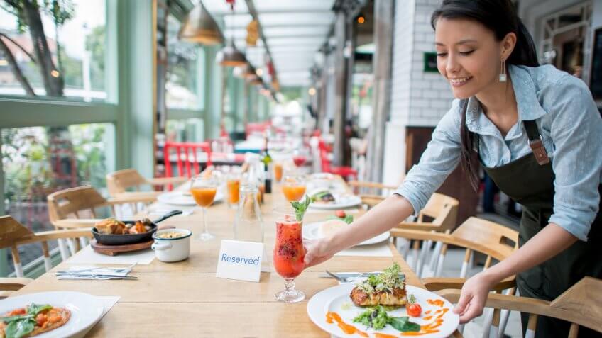 Happy Latin American waitress working at a restaurant and serving dinner on the table.