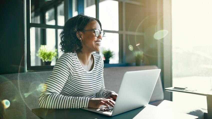 Smiling young African woman wearing glasses looking out of a window while sitting at a table working online with a laptop.