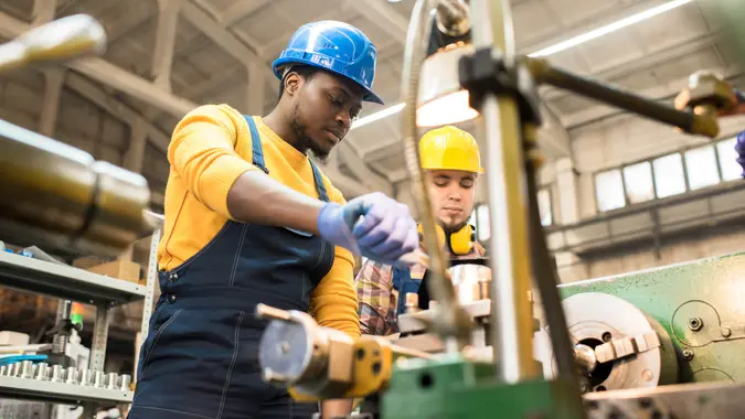 Multi-ethnic team of workers wearing overalls and protective helmets using lathe in order to machine workpiece, interior of spacious production department on background.