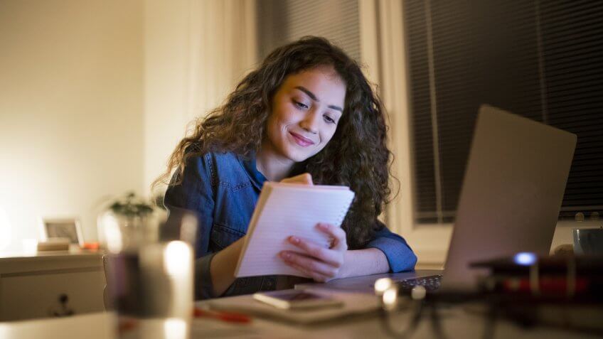 Young beautiful woman sitting at desk late at night, laptop in front of her, writing something into notebook.