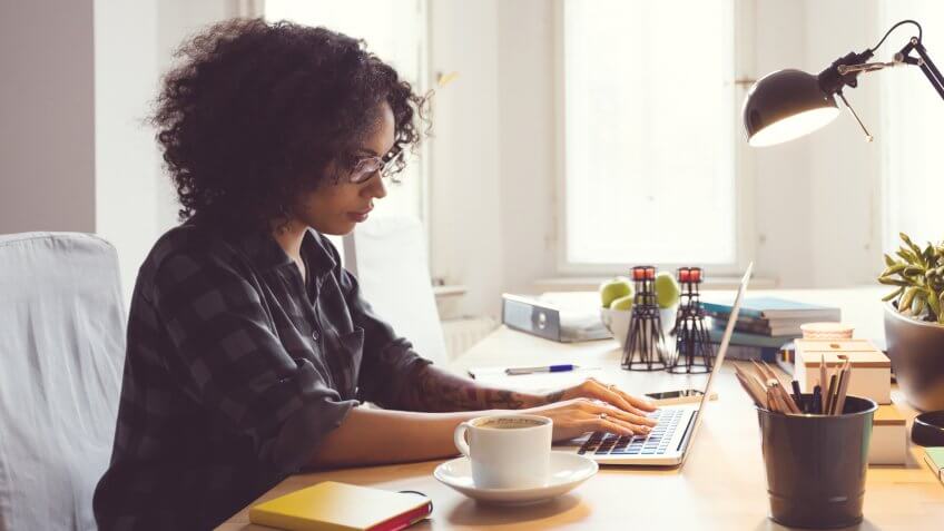 Afro young woman sitting at the table in a home office, using laptop.
